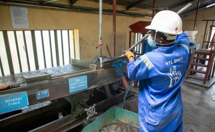 Trabalhador usando uniforme azul opera uma mesa vibratória para processamento de minério em uma unidade industrial.