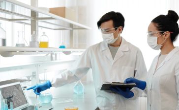 Dois pesquisadores usando jalecos analisam dados em um computador dentro de um laboratório.
