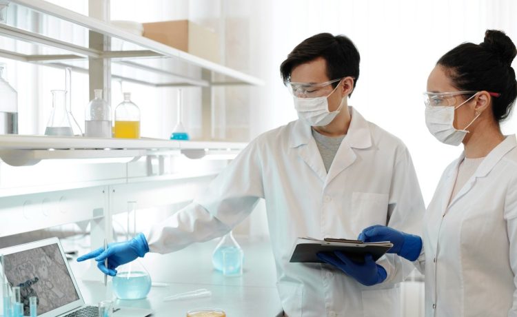 Dois pesquisadores usando jalecos analisam dados em um computador dentro de um laboratório.