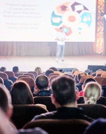 Público acompanha atentamente uma apresentação em um auditório iluminado por luzes azuis.