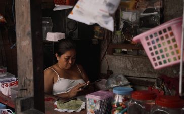 Mulher usa o celular dentro de um pequeno comércio repleto de produtos.