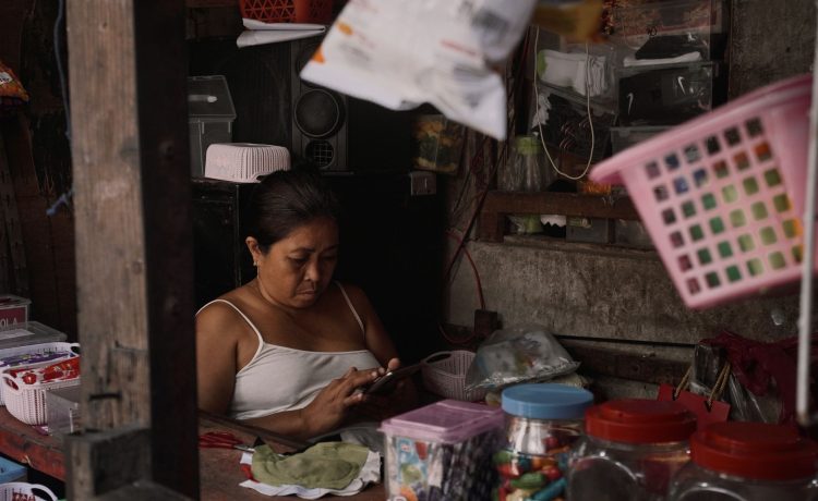 Mulher usa o celular dentro de um pequeno comércio repleto de produtos.